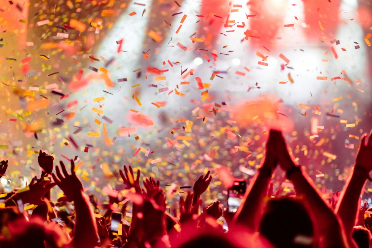 Rear view of a large group of people in front of a music festival stage. Crowd is excited and dancing, raising hands, clapping, punching the air, filming with mobile phones, etc...