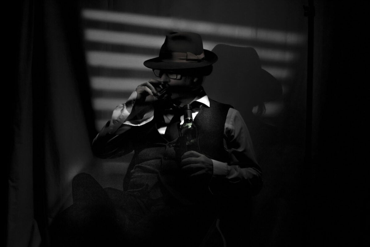Film Noir style detective drinking and wearing a Fedora hat, tie and waistcoat in a dark, seedy office with a stylized Venetian blind window behind. The face is partially obscured by the hat brim and shadow.